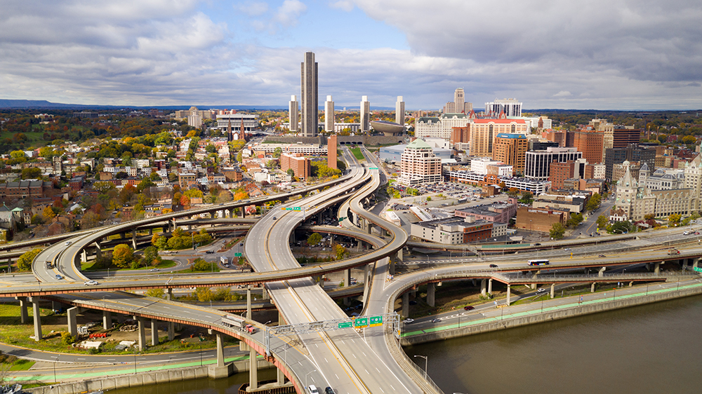 An aerial photo of Albany, New York, shows freeways on and off ramps leading to the downtown core featuring multi-story buildings on the right, a residential neighborhood on the left, and an industrial area in the background.
