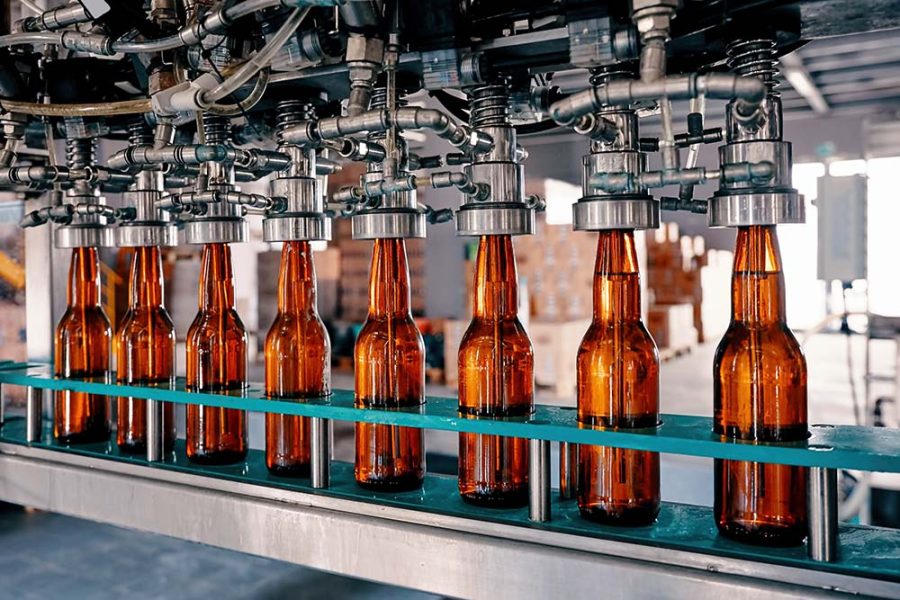 A row of translucent brown beer bottles rides on a conveyor belt in a brewery while being filled and capped by automated machinery