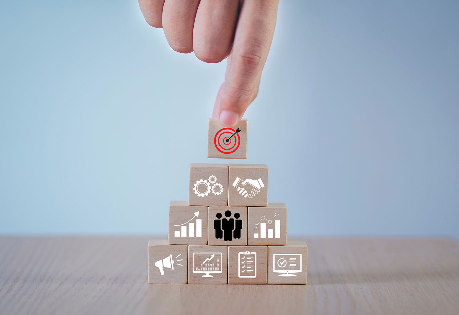 A hand stacks small wooden blocks with different icons on them representing aspects of successful prospecting and lead generation, including marketing, data analysis, making connections, and in-person meetings.