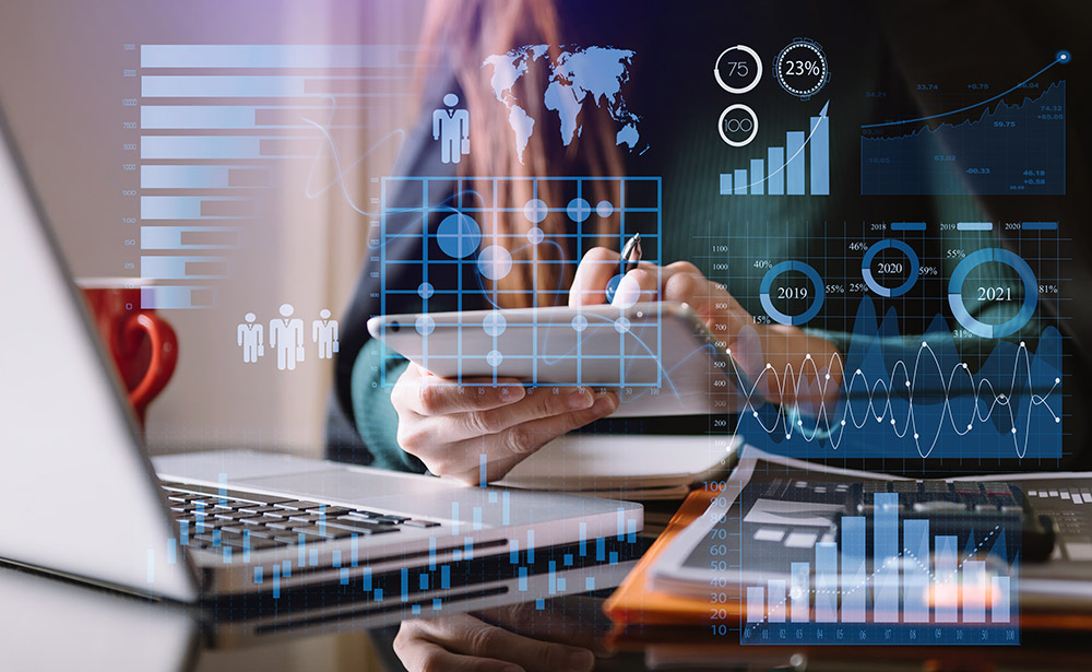 A woman sits at a desk with a laptop computer open in front of her while she uses a tablet computer. Multiple images of different industry data charts and graphs are superimposed over the top of the image.
