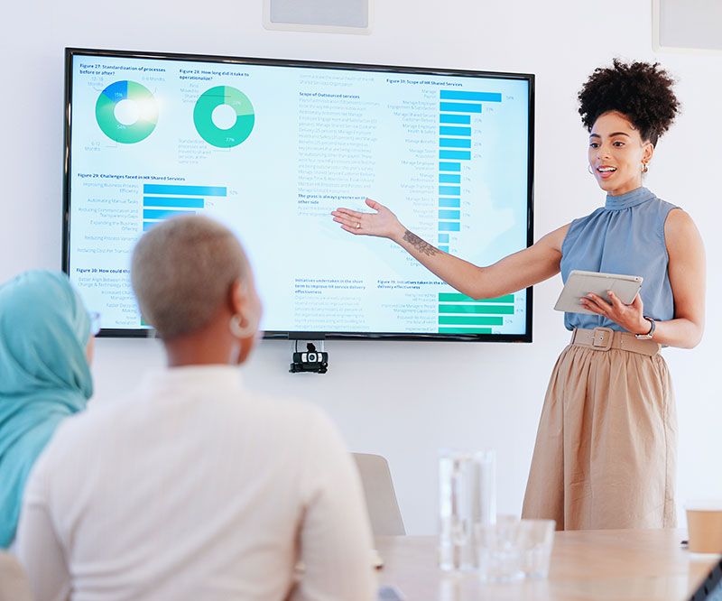 A woman uses data presented on a large video monitor to tell the story about an organization to an audience