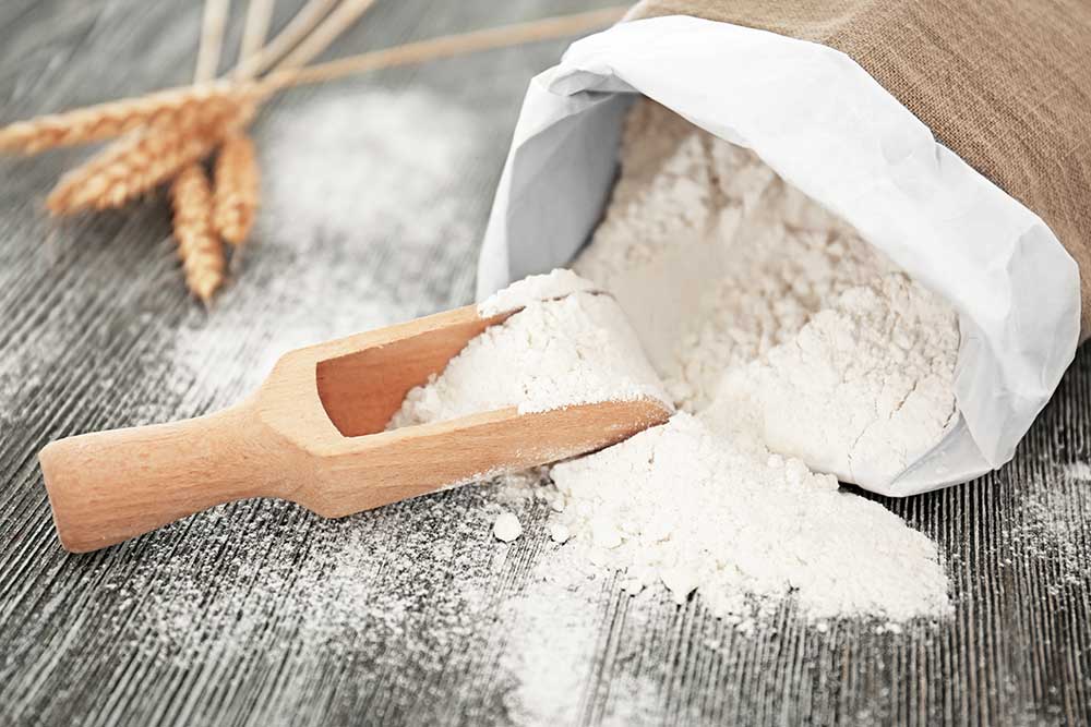 A bag of wheat flour sits open on a countertop with a wooden scoop and a few stalks of wheat next to it