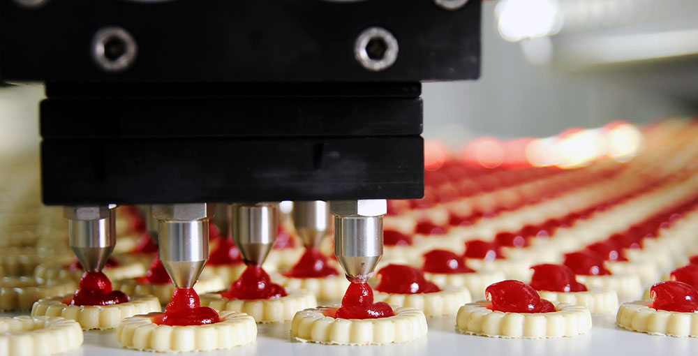 A machine adds red jelly to the top of many sugar cookies on a conveyor belt in a food production factory