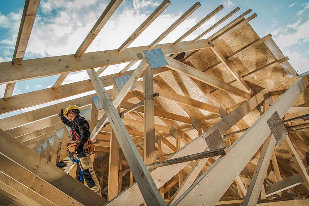 A worker wearing a hard hat and tool belt holds a hammer while standing within the framing beams of a new house under construction