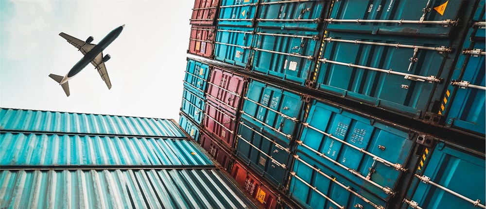 A commercial jet flies over tall stacks of larger cargo containers