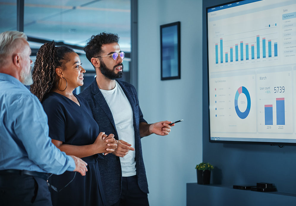 Data is presented by a consultant to two stakeholders on a large video monitor