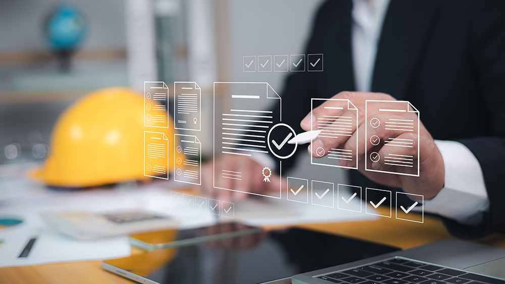 A man in a suit sits at a desk with blueprints, a yellow hard hat, a tablet computer, and a smartphone. He uses a digital pen to review approved real estate development plans.