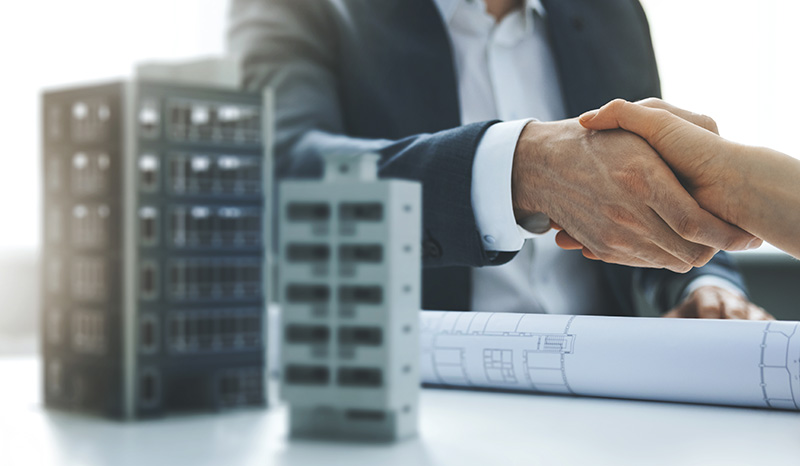 A real estate developer shakes hands with an investor over rolled development construction plans and small models of what the proposed buildings would look like