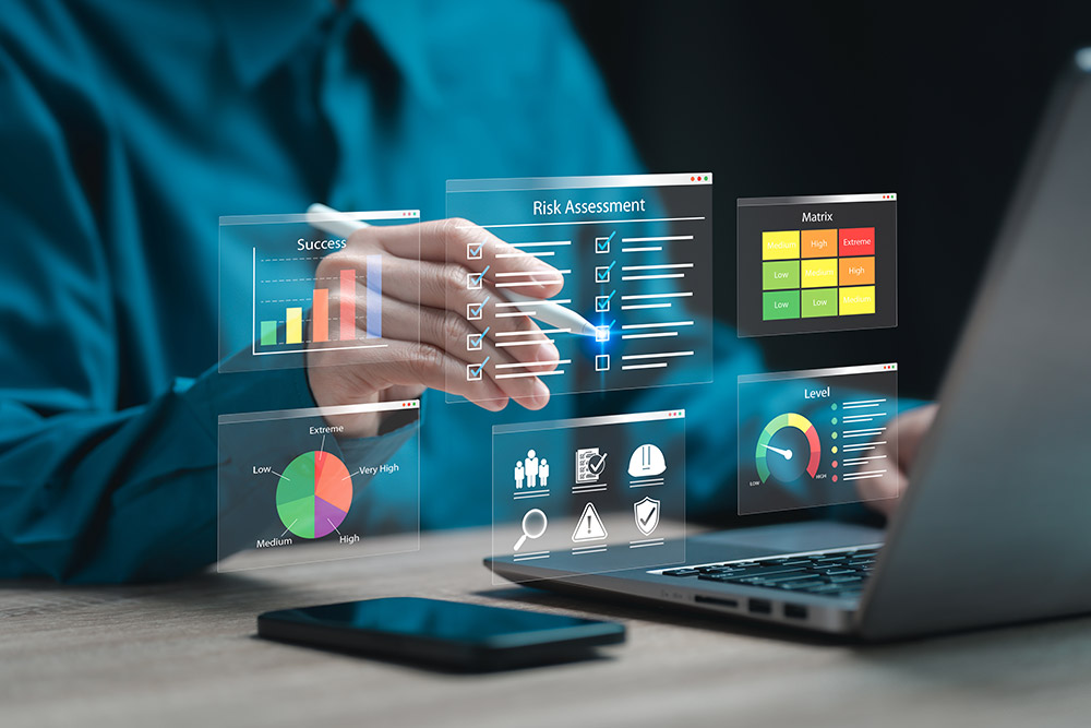 A person in a blue long-sleeves dress shirt sits in front of an open laptop computer and uses a digital pen to check items off a Risk Assessment list. Additional risk assessment-related data and charts also appear.