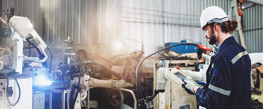 A robotics engineer in a white hard hat operates a white robotic arm in a manufacturing factory full of machinery and equipment
