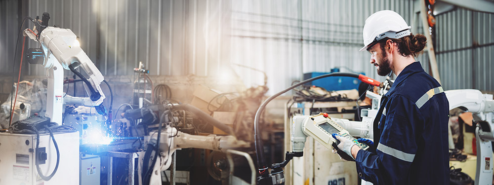 A robotics engineer in a white hard hat operates a robotic arm that is completing a welding task in a manufacturing factory full of machinery and equipment