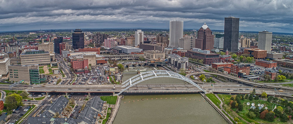 An aerial view of Rochester, New York, shows the Genesee River, the Frederick Douglass Susan B. Anthony Bridge, Court Street Dam, and many skyscrapers and smaller commercial and residential buildings