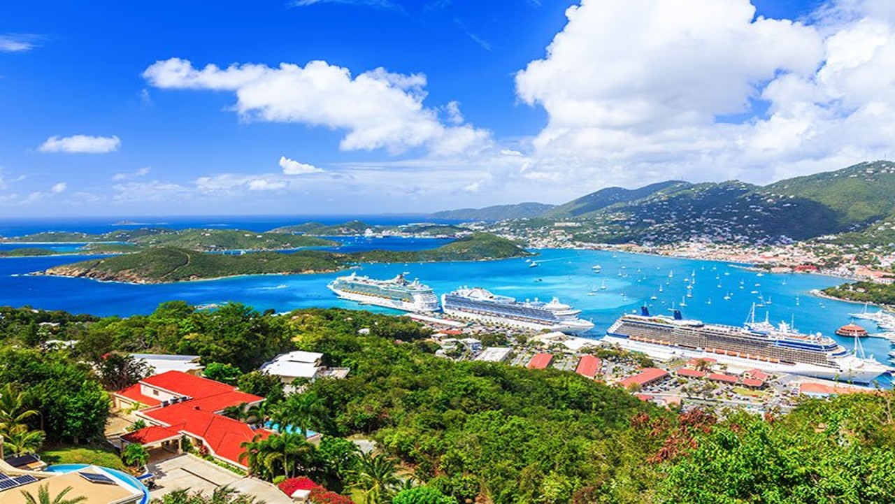 Hillside view of cruise ships docked in a cove in the US Virgin Islands with lots of green forested areas, bright blue water, and a few buildings in the foreground
