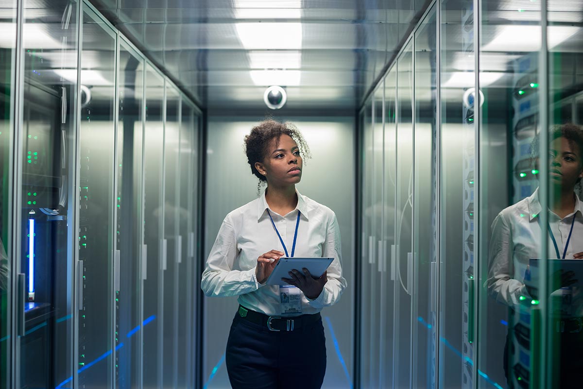 A woman holding a tablet computer walks between two rows of computer servers in a data center